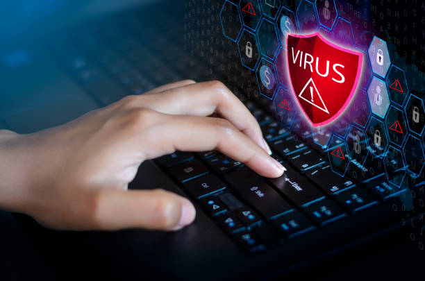 Close-up of a woman's hand a laptop keyboard. On the screen is a bright red shield icon with an exclamation point and the word "VIRUS"